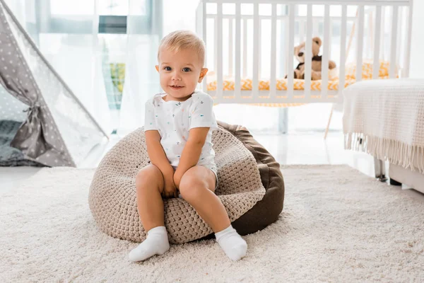 Entzückend lächelndes Kleinkind sitzt auf Bohnensackstuhl und blickt in Kinderzimmer in die Kamera — Stockfoto