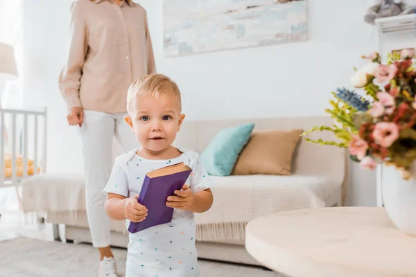 Entzückendes Kleinkind hält Buch in der Hand und schaut in die Kamera drinnen mit Mutter im Hintergrund — Stockfoto