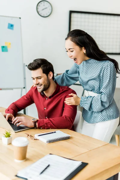 Colegas multiétnicos emocionados animando y utilizando el ordenador portátil en la oficina - foto de stock