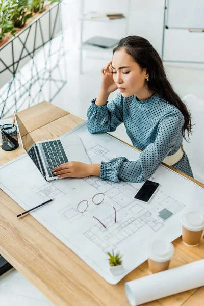 Estressado asiático feminino arquiteto tendo dor de cabeça enquanto usando laptop no escritório — Fotografia de Stock