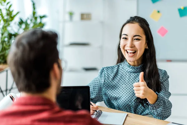 Smiling asian businesswoman sitting and showing thumb up sign to colleague in office — Stock Photo