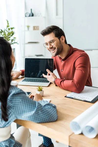 Arquitecto masculino sentado en la mesa y apuntando con el dedo a la computadora portátil con pantalla en blanco en la oficina - foto de stock