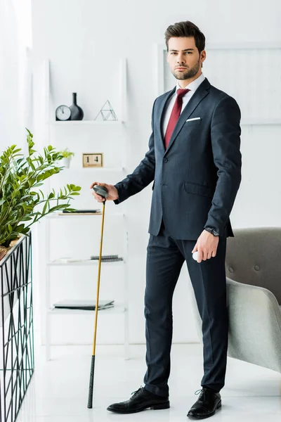 Businessman in formal wear holding golf club with golf ball and looking at camera in office — Stock Photo