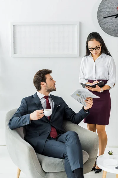 Mujer de negocios asiática escribiendo en cuaderno mientras hombre de negocios sentado en sillón con periódico y café en la oficina - foto de stock