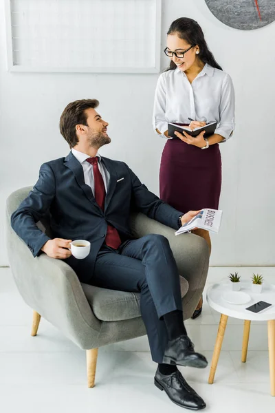 Mujer de negocios asiática escribiendo en cuaderno mientras hombre de negocios sentado en sillón con periódico y café en la oficina - foto de stock