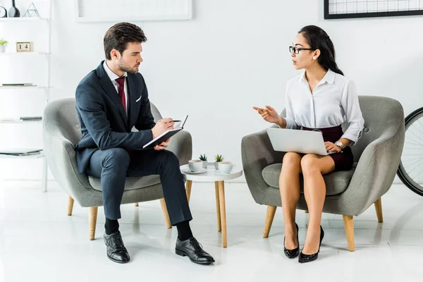 Multiethnic businesspeople with notebook and laptop talking while sitting in waiting hall — Stock Photo