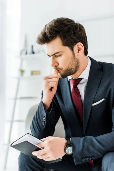 Hombre de negocios pensativo en ropa formal sentado y la celebración de cuaderno en la oficina - foto de stock