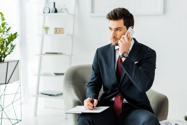 Schöner Geschäftsmann in formeller Kleidung, der auf einem Sessel sitzt und in einem Notizbuch witzelt, während er im Büro mit dem Smartphone spricht — Stockfoto