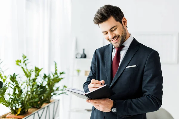 Красивый улыбающийся бизнесмен в формальной одежде пишет в блокноте в офисе — стоковое фото