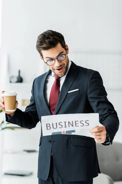 Surprised businessman in formal wear holding coffee to go and reading newspaper in office — Stock Photo