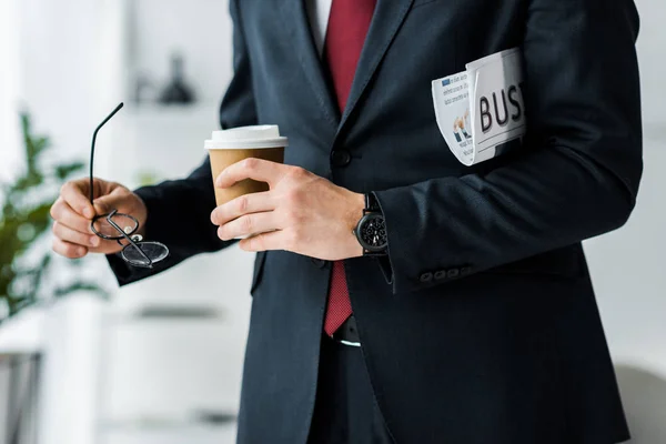 Cropped view of businessman in formal wear holding coffee to go and newspaper in office — Stock Photo