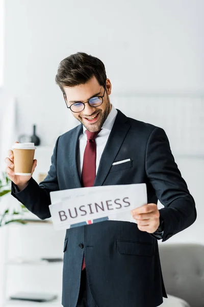 Homem de negócios sorridente em desgaste formal segurando café para ir e ler jornal no escritório — Fotografia de Stock