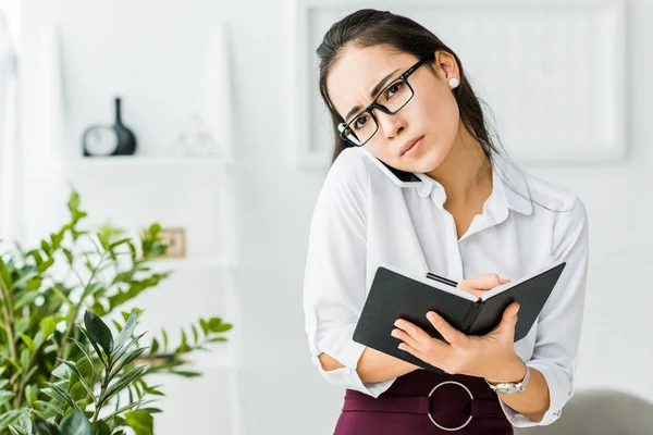 Asian businesswoman talking on smartphone and writing in notebook in office — Stock Photo