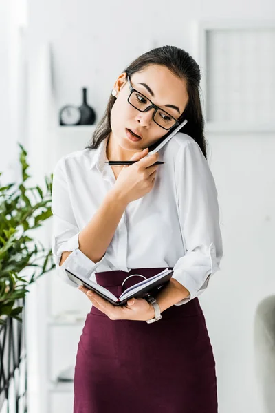 Asian businesswoman talking on smartphone while holding notebook in office — Stock Photo