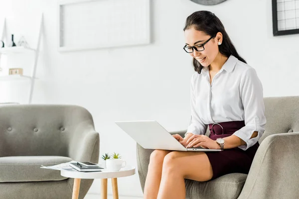 Красива усміхнена азіатська бізнес-леді в окулярах сидить на кріслі і використовує ноутбук в офісі — стокове фото