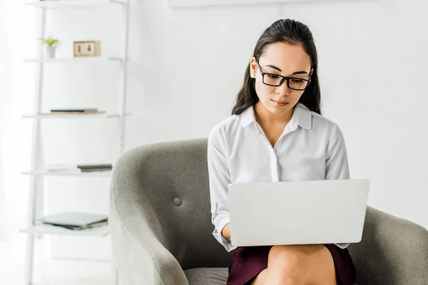 Hermosa asiático mujer de negocios en gafas sentado en sillón y el uso de portátil en la oficina — Stock Photo