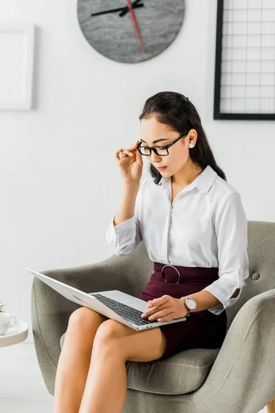 Belle asiatique femme d'affaires dans des lunettes assis sur fauteuil et en utilisant ordinateur portable au bureau — Photo de stock