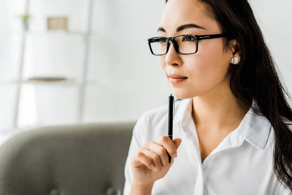 Beautiful asian businesswoman in formal wear and glasses holding pen and looking away in office — Stock Photo