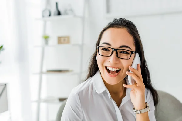 Cheerful asian businesswoman in glasses talking on smartphone in office — Stock Photo