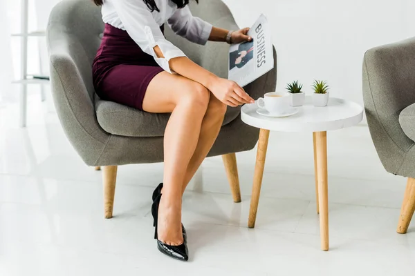 Vista ritagliata di donna d'affari seduta sulla poltrona e che tiene il giornale con la tazza di caffè in ufficio — Foto stock