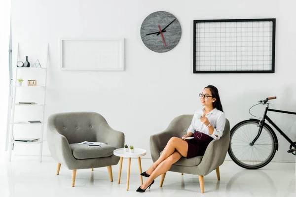 Sonriente asiático mujer de negocios sentado en sillón con café taza en moderno oficina - foto de stock