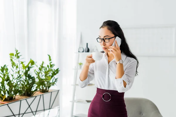 Hermosa seria asiático mujer de negocios hablando en smartphone mientras teniendo café descanso en oficina - foto de stock