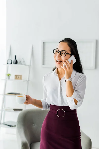 Beautiful laughing asian businesswoman talking on smartphone while having coffee break in office — Stock Photo