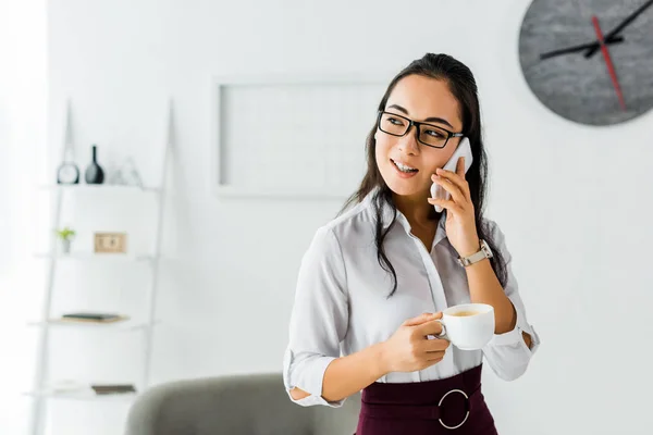 Beautiful smiling asian businesswoman talking on smartphone while having coffee break in office — Stock Photo
