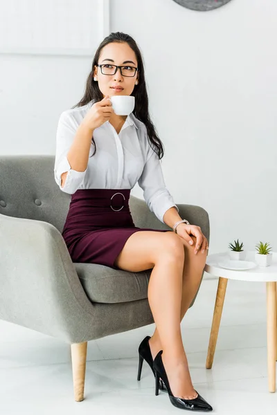 Bella asiatico businesswoman in occhiali seduta su poltrona e avendo caffè pausa in ufficio — Foto stock
