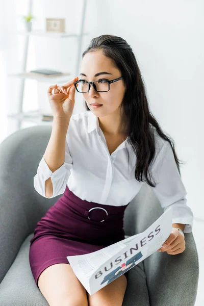 Beautiful asian businesswoman holding glasses and newspaper while sitting on armchair in office — Stock Photo