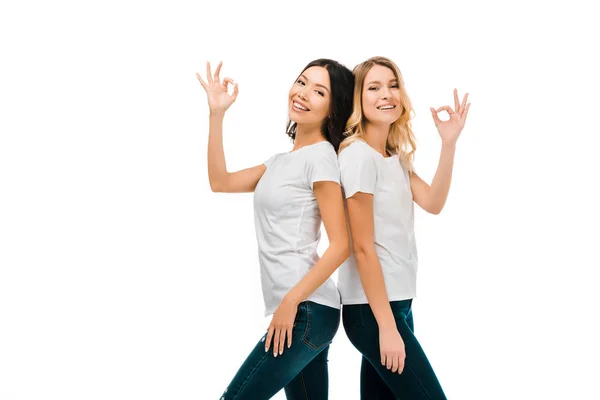 Feliz jóvenes mujeres de pie espalda con espalda y mostrando ok signo aislado en blanco - foto de stock