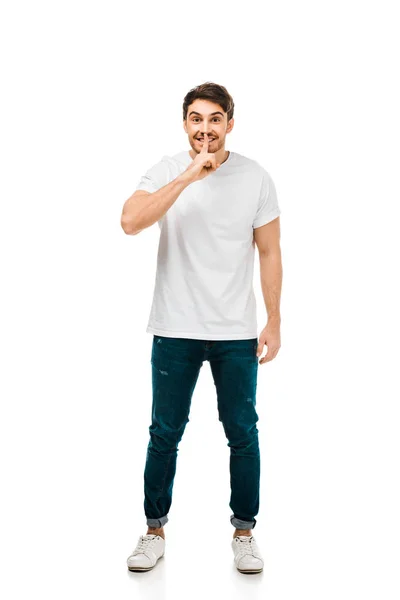 Full length view of man in white t-shirt gesturing for silence with finger on lips and looking at camera isolated on white — Stock Photo