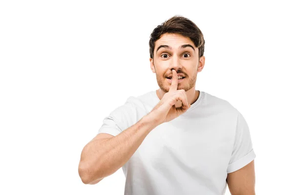 Jeune homme en t-shirt blanc gesticulant pour le silence avec le doigt sur les lèvres et regardant la caméra isolée sur blanc — Photo de stock