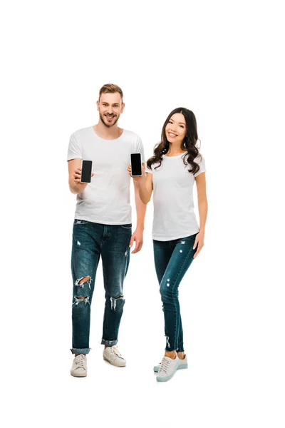 Feliz joven pareja sosteniendo teléfonos inteligentes con pantallas en blanco y sonriendo a la cámara aislada en blanco - foto de stock