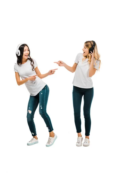 Belles filles heureuses dans les écouteurs dansant et pointant avec les doigts isolés sur blanc — Photo de stock