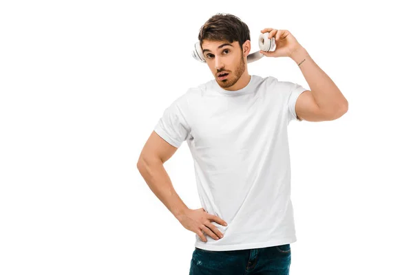 Joven con la boca abierta sosteniendo los auriculares y mirando a la cámara aislada en blanco - foto de stock