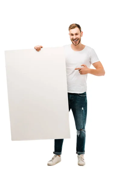 Красивый молодой человек, указывающий пальцем на пустой плакат и улыбающийся в камеру, изолированную на белом — стоковое фото
