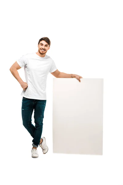 Повний вигляд красивого молодого чоловіка, що стоїть з порожнім плакатом і посміхається на камеру ізольовано на білому — стокове фото