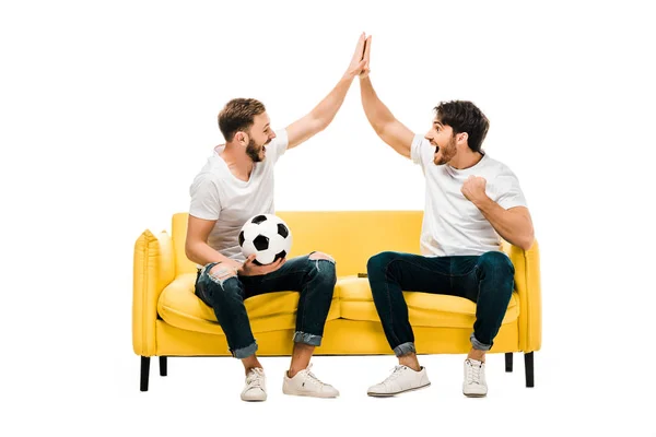 Feliz joven sentado en el sofá con pelota de fútbol y dando cinco alto aislado en blanco - foto de stock