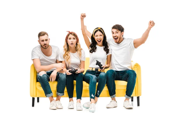 Emotional young friends sitting on sofa and playing with joysticks isolated on white — Stock Photo