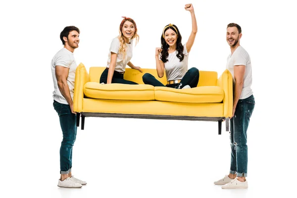 Allegri giovani uomini che trasportano divano con belle ragazze sedute su di esso isolate su bianco — Foto stock