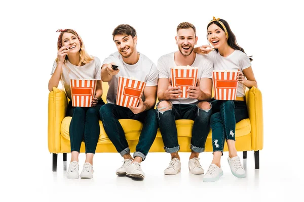 Glückliche junge Freunde, die Popcornboxen in der Hand halten und fernsehen, während sie auf dem gelben Sofa sitzen, isoliert auf weiß — Stockfoto