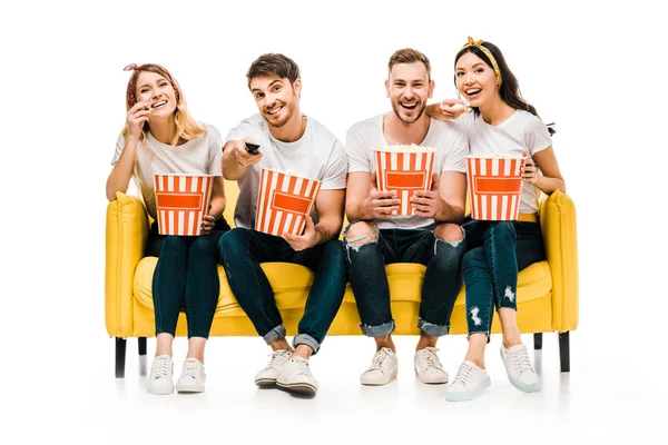 Heureux jeunes amis tenant boîtes de maïs soufflé et regarder la télévision tout en étant assis sur le canapé jaune isolé sur blanc — Photo de stock