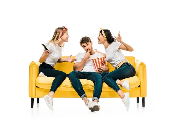 Sorrindo jovens mulheres falando e homem comendo pipocas enquanto sentados juntos no sofá isolado no branco — Fotografia de Stock