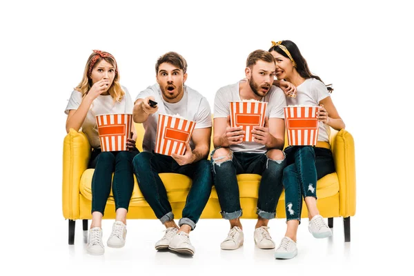 Shocked young friends eating popcorn from boxes and watching tv while sitting on yellow sofa isolated on white — Stock Photo