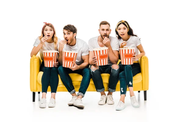 Молодые друзья едят попкорн из коробок и смотрят в камеру, сидя на желтом диване, изолированном на белом — стоковое фото