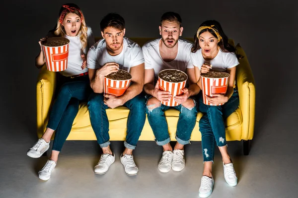 Vista ad angolo alto di giovani amici scioccati che tengono scatole di popcorn e guardano la TV seduti insieme sul divano sul grigio — Foto stock