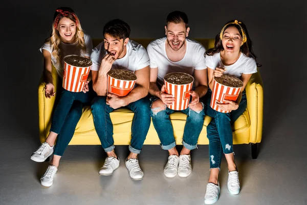 Vista ad alto angolo di felici giovani amici seduti sul divano e mangiare popcorn da scatole su grigio — Foto stock