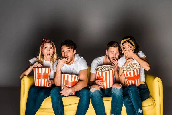 Jóvenes amigos asustados comiendo palomitas de maíz y viendo la televisión mientras están sentados juntos en el sofá en gris - foto de stock