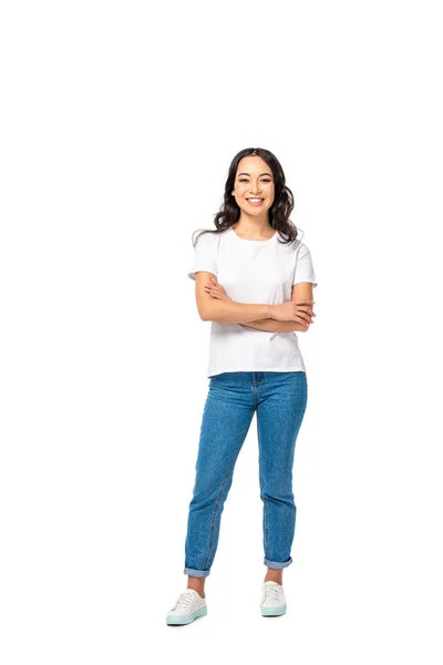 Улыбающаяся азиатская молодая женщина в белой футболке и синих джинсах, стоящих со скрещенными руками, изолированными на белом — стоковое фото